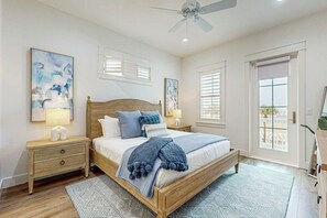 A bedroom with a wooden bed, two bedside tables, a ceiling fan, and a large window with blinds. - Grand Rising | Gulf Views | Private Elevator (Port Aransas)