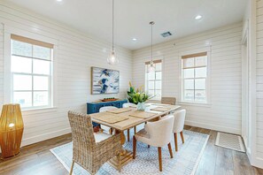 A dining area with a long table, wicker chairs, a vase with plants, and artwork on the wall. - Grand Rising | Gulf Views | Private Elevator (Port Aransas)