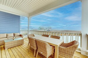 Área externa coberta com deck de madeira, móveis de vime e vista para casas sobre a água.