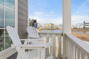 En terrass med vita Adirondack-stolar med utsikt över en sjö eller ett annat vattendrag och en rad hus.