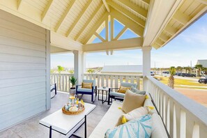 A covered outdoor seating area with a white railing, a table with a basket of food, and a sofa with cushions. - Ocean Pearl | Gulf Front | Steps to Beach (Port Aransas)