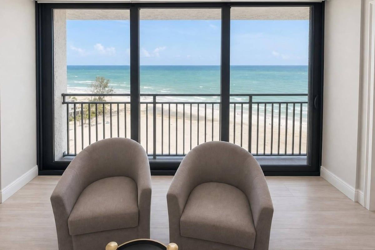 Ein Zimmer mit zwei Sesseln, die zu einem großen Fenster mit Blick auf einen Strand blicken.