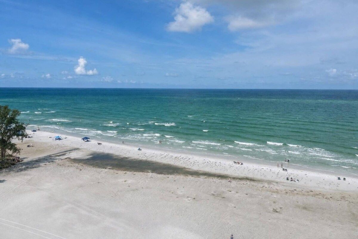 A beach with clear blue water, sandy shore, and a few umbrellas.