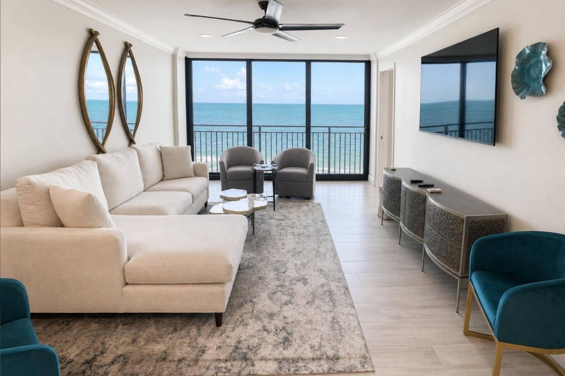 A living room with a large window overlooking the ocean, a ceiling fan, and a flat-screen TV mounted on the wall.