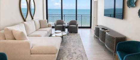 A living room with a large window overlooking the ocean, a ceiling fan, and a flat-screen TV mounted on the wall.