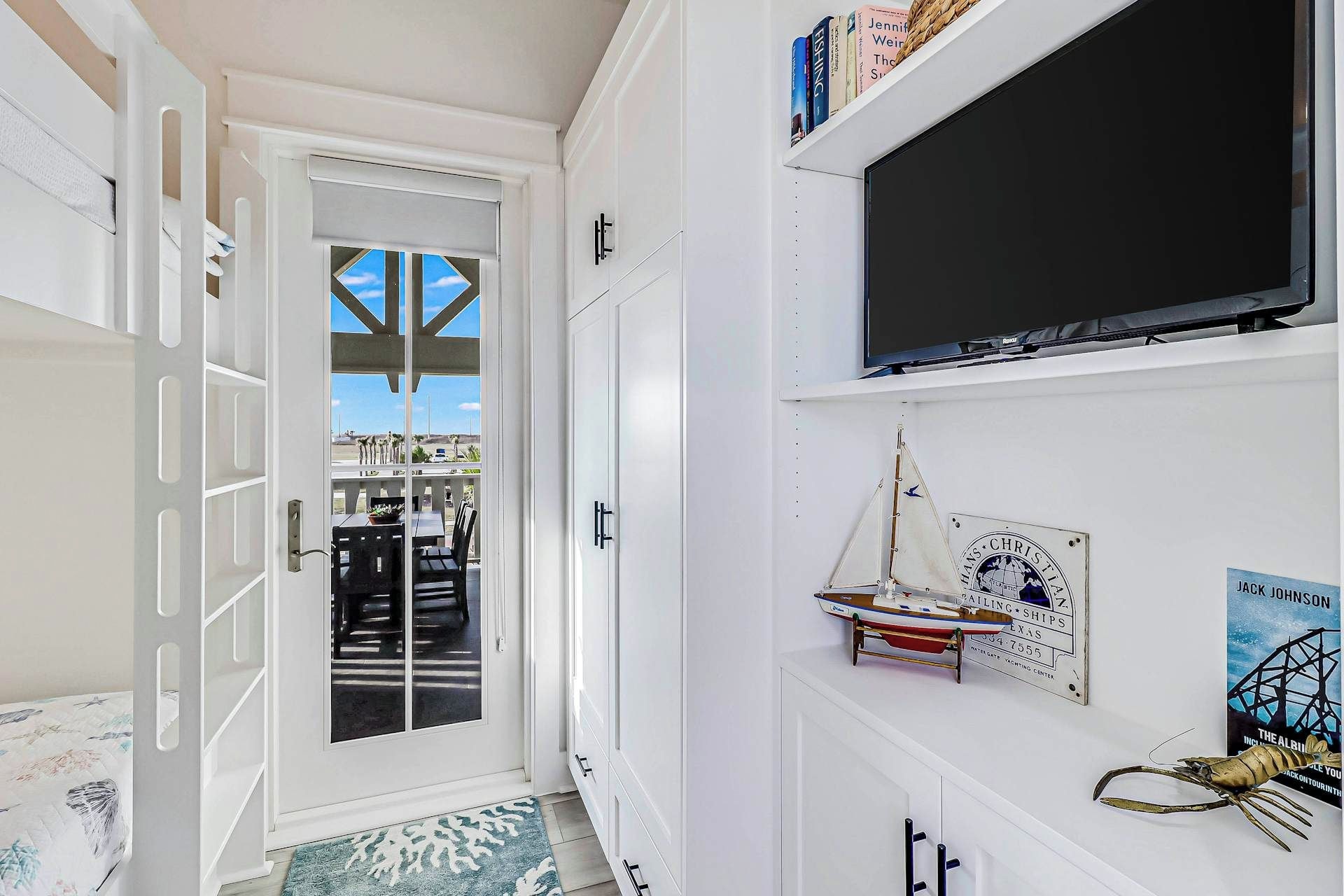 Een hal met een deur die uitkomt op een balkon, een wandtelevisie en een klein model zeilboot aan de muur.