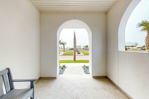 A corridor with a bench, leading to an outdoor area with a palm tree and a view of a parking lot. - Mimosa Manor | Steps to Beach | Resort Amenities (Port Aransas)