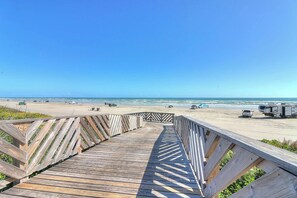 Ein hölzerner Steg führt zu einem Strand mit Fahrzeugen und blauem Himmel.