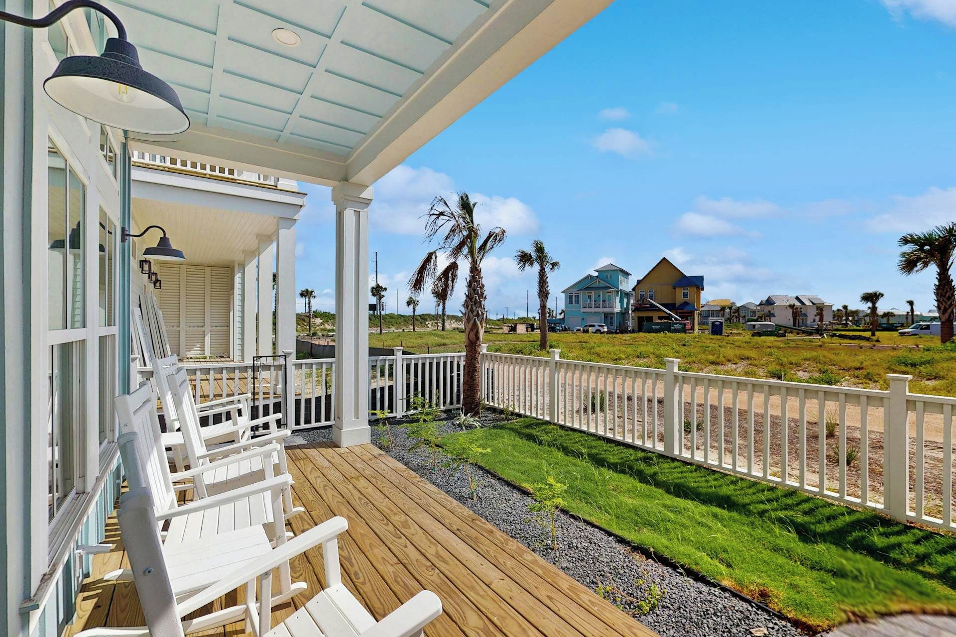 A wooden deck with white chairs, a covered porch with a lamp, and a view of houses and palm trees.