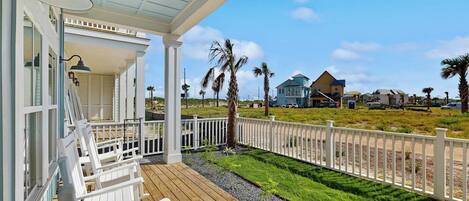 A wooden deck with white chairs, a covered porch with a lamp, and a view of houses and palm trees.