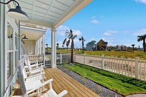 A wooden deck with white chairs, a covered porch with a lamp, and a view of houses and palm trees. - Ocean Pearl | Pet Friendly | Steps to Beach (Port Aransas)