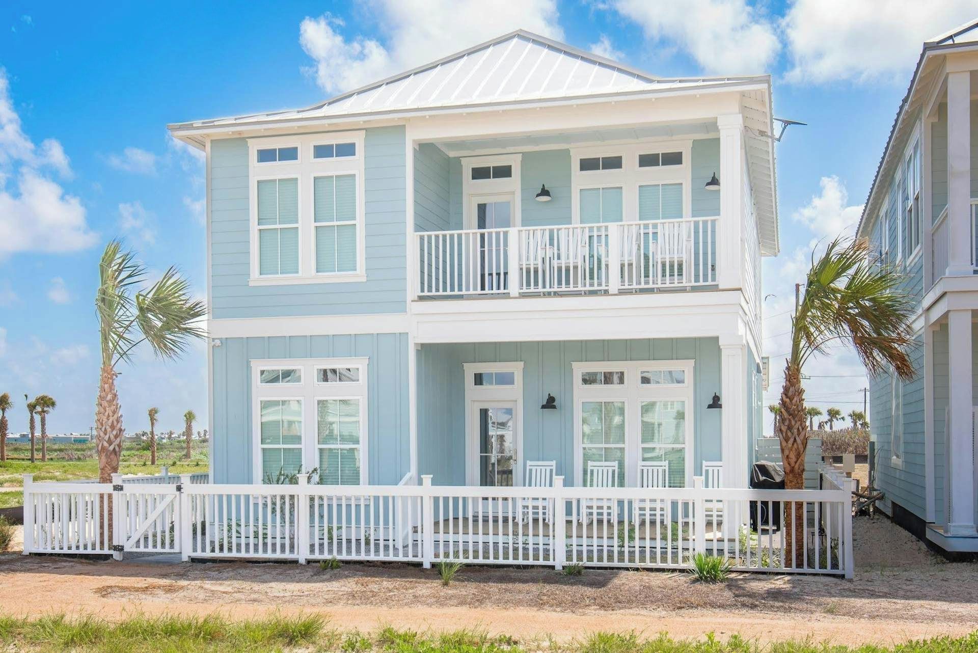 Una casa fronte spiaggia su due piani, con un recinto bianco, diverse finestre e un balcone.