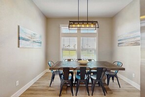 A dining area with a wooden table, six chairs, a hanging light fixture, and two windows with blinds. - Ocean Pearl | Pet Friendly | Steps to Beach (Port Aransas)