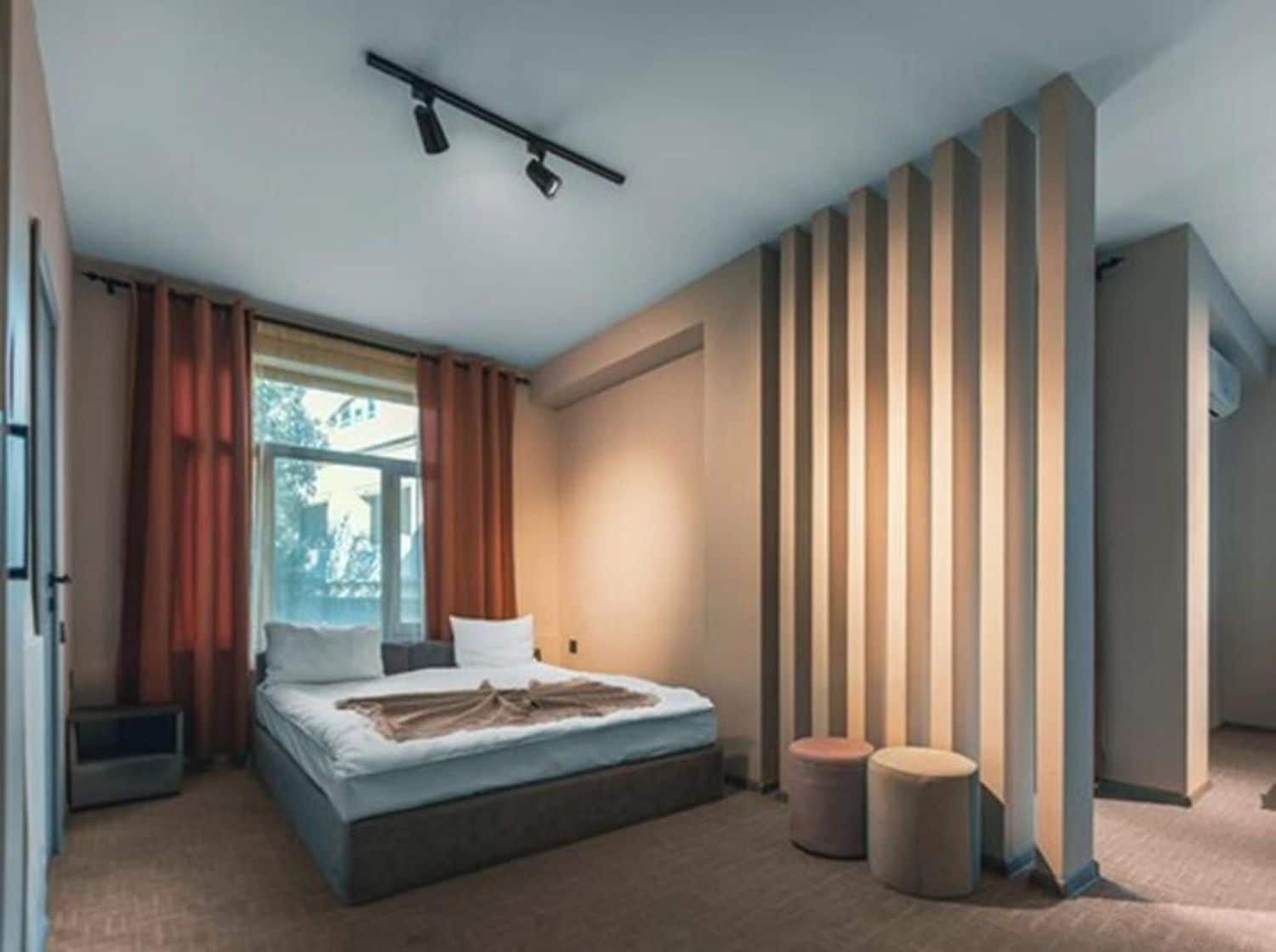 Een moderne slaapkamer met een bed, een raam met gordijnen, een wand met verticale lijnen en twee poefs.