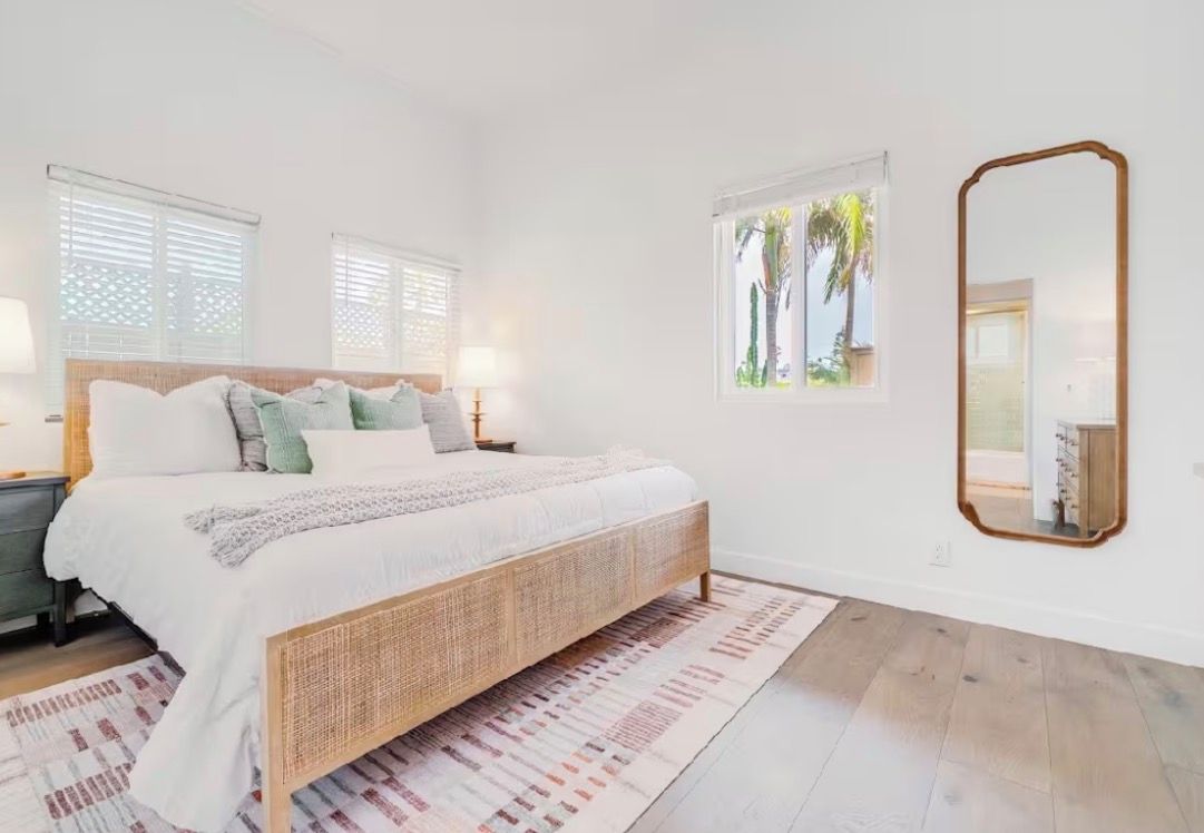 A bedroom with a large bed, a nightstand, a mirror, and a window with a view of palm trees.