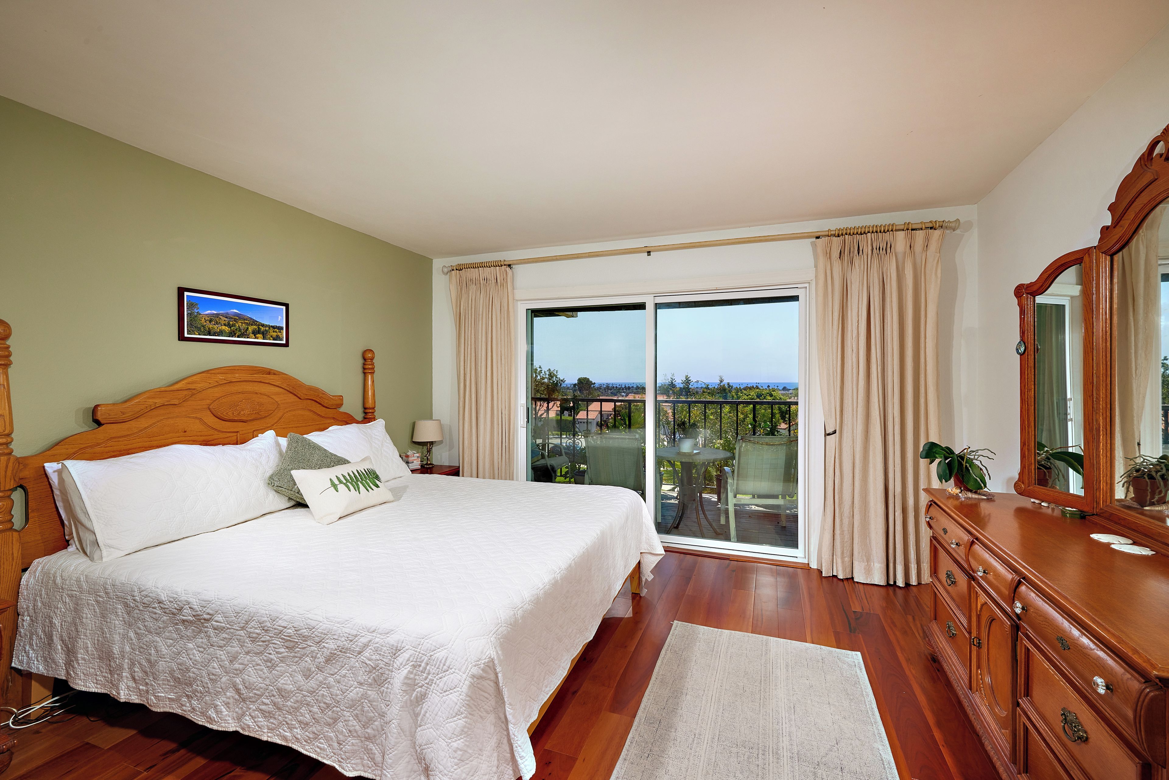 A bedroom with a large bed, a wooden dresser, a mirror, and a view through a glass door to the outside.