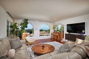 A spacious living room with a sofa, armchair, coffee table, piano, and a large TV. - Beautiful 2-bedroom condo in Carlsbad near the beach, with pool, and ocean view! (Carlsbad)