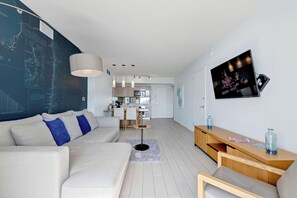 A modern living room with a sofa, a coffee table, and a TV mounted on the wall. - Modern 2-BR APT - Condo Near Ocean w Amenities (Hallandale Beach)