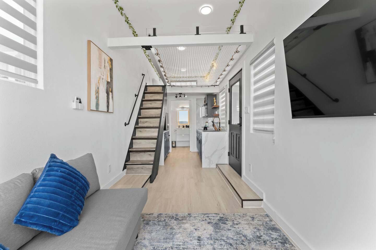A modern interior with a staircase leading to a kitchen area, a grey sofa with a blue cushion, and a patterned rug.