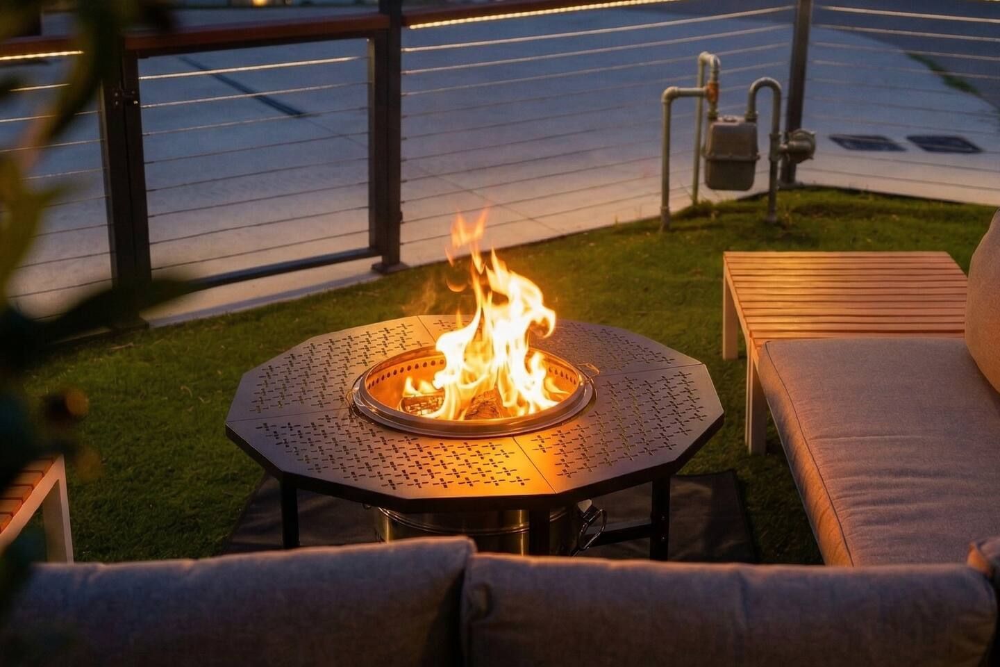A round outdoor fire pit with flames, surrounded by a metal table and two cushioned chairs.