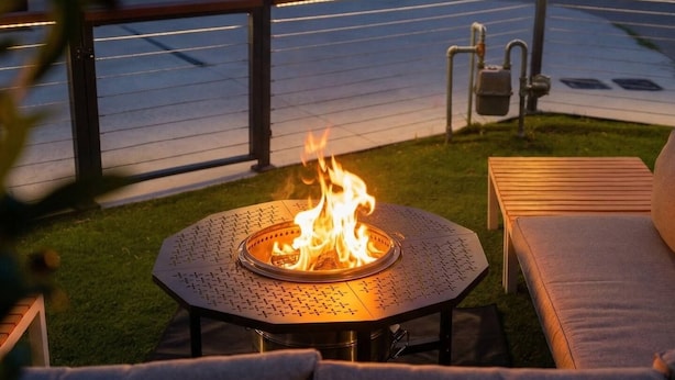 En rund udendørs pejsebrænding med flammer, omgivet af et metalbord og to polstrede stole.
