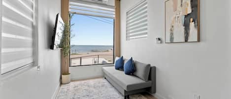 A modern living room with a grey sofa, blue cushions, a large window with a view of the sea, and a painting on the wall.