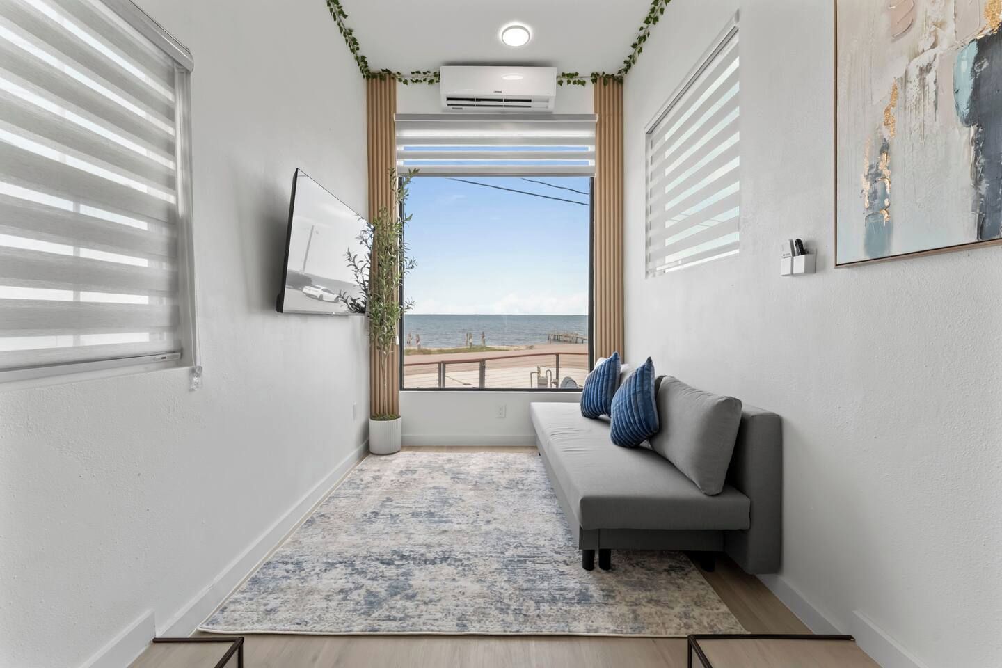Ein modernes Wohnzimmer mit einem grauen Sofa, einem großen Fenster mit Meerblick und einer Wand-Klimaanlage.
