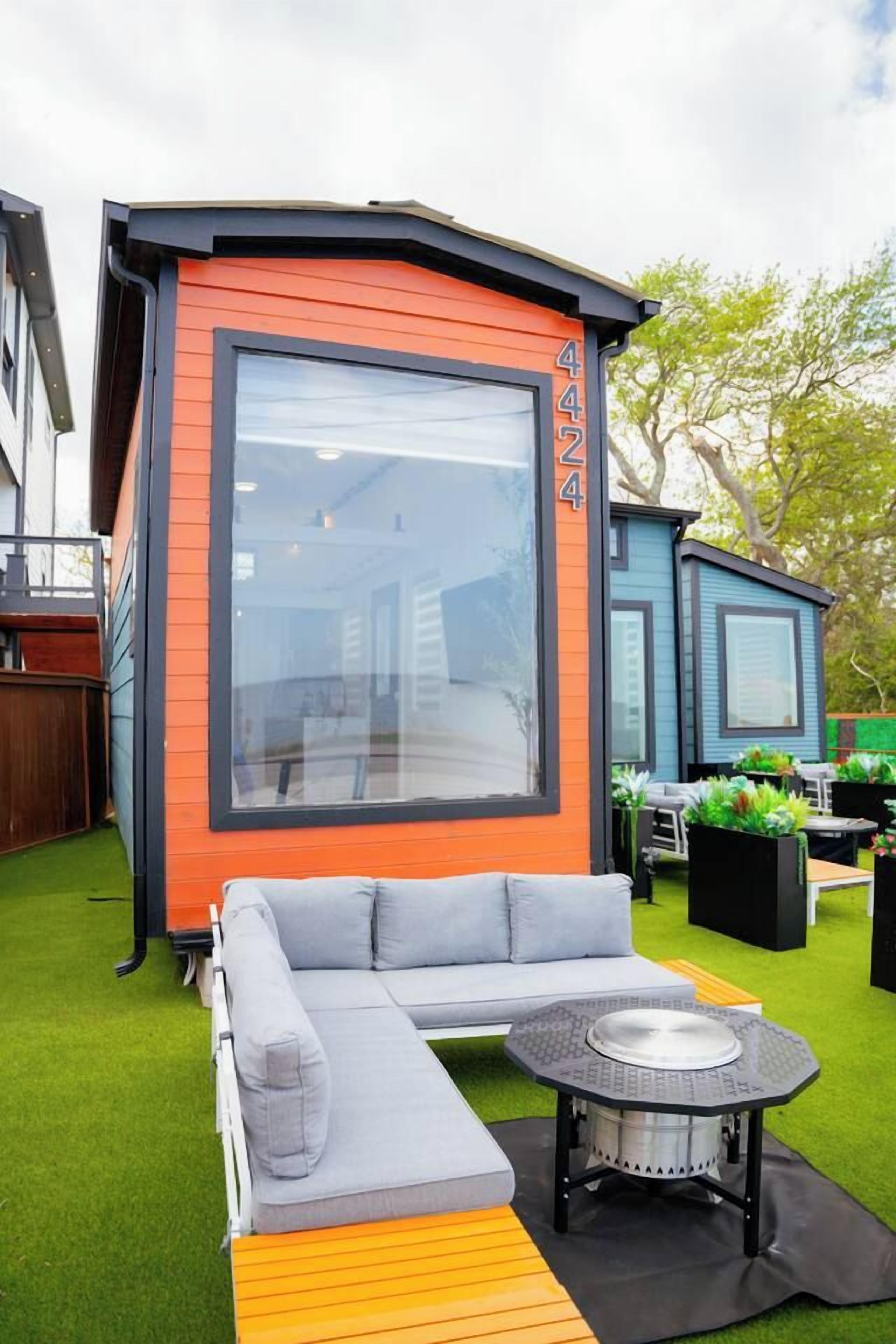 A compact, orange and blue modular building with a large window, a grey sofa, and a round table with a metal top.