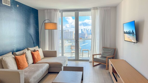 Una sala de estar moderna con un sofá, una butaca, una mesa de centro, un televisor y vistas a un paisaje urbano a través de un ventanal grande.