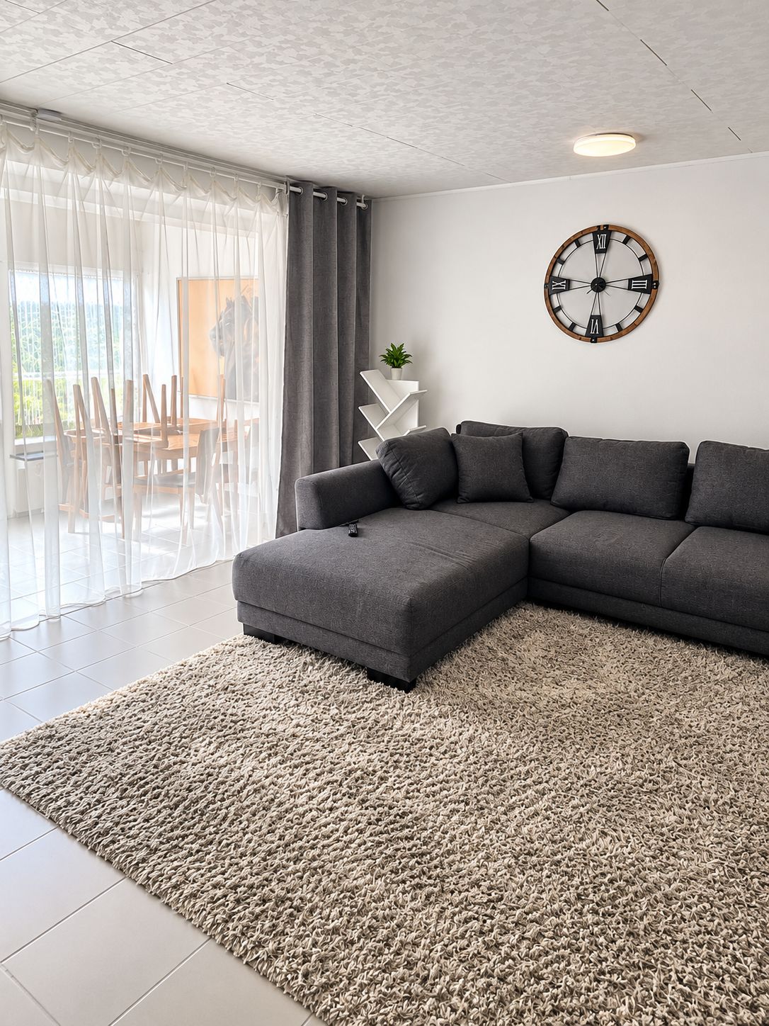 Una sala moderna con un sofá modular, un reloj de pared redondo y vista a un comedor a través de cortinas traslúcidas.