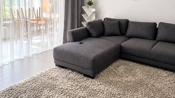 Una sala moderna con un sofá modular, un reloj de pared redondo y vista a un comedor a través de cortinas traslúcidas.