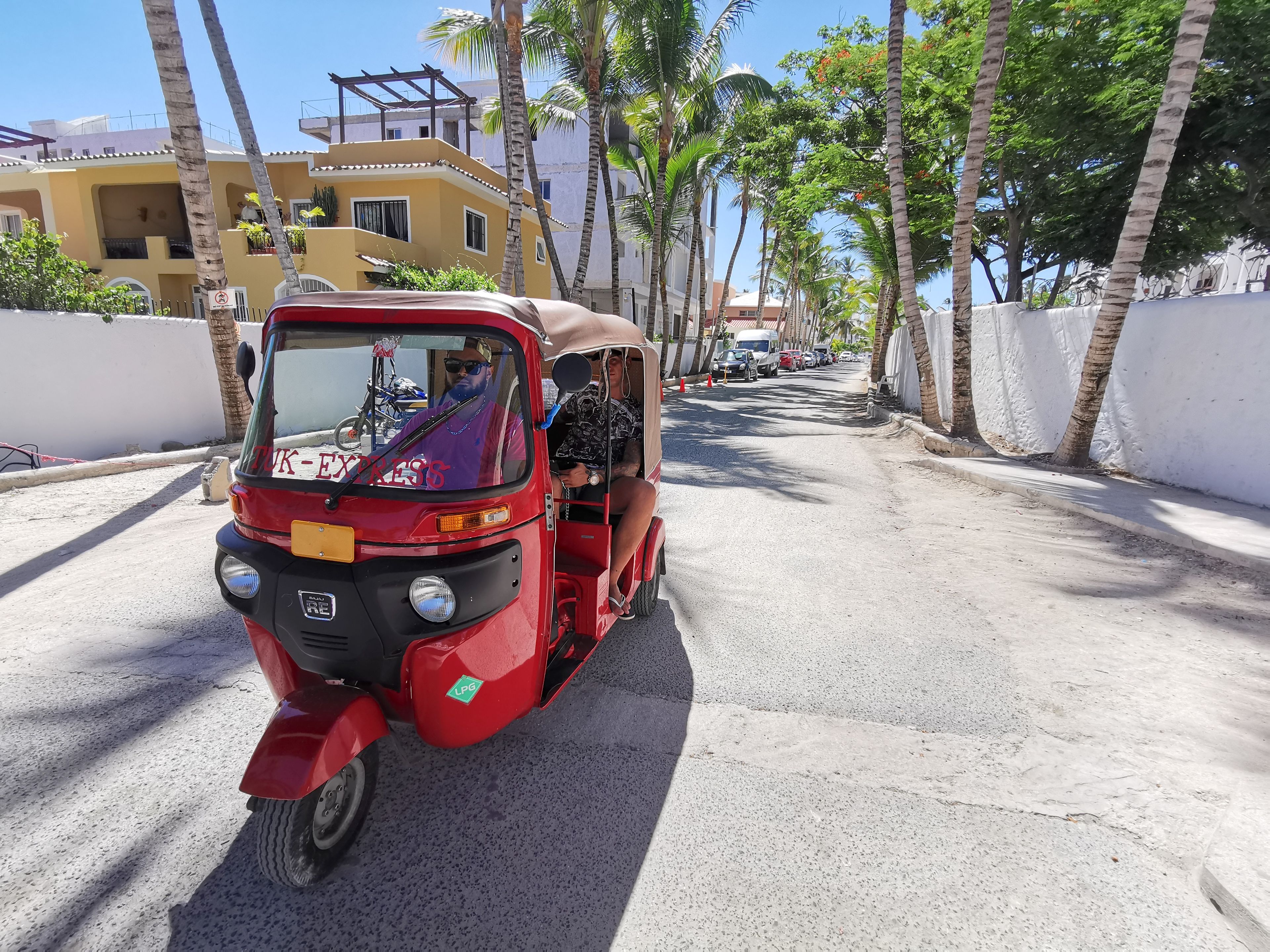 Un rickshaw rojo circulando por una calle con palmeras y edificios al fondo.