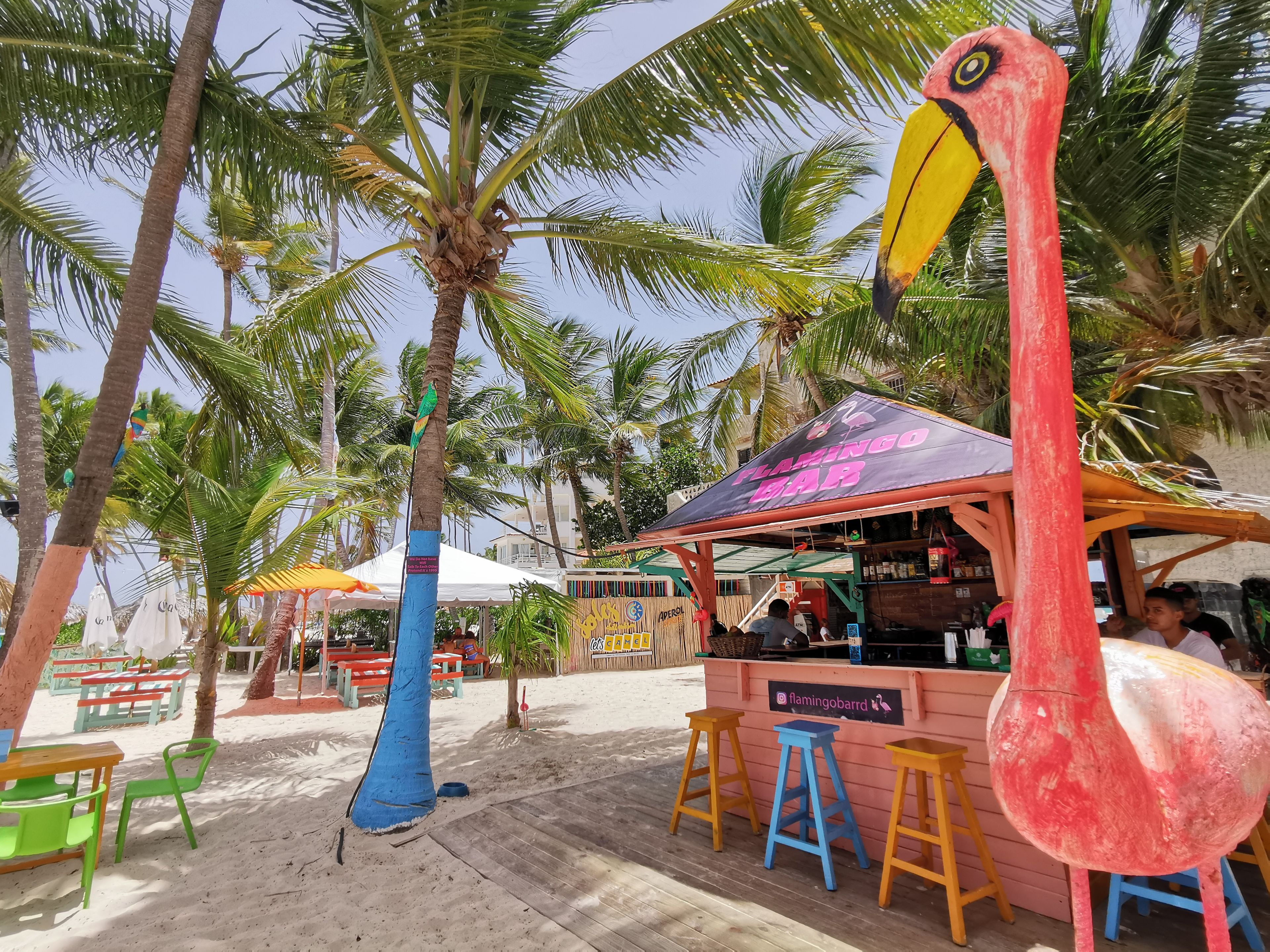 A beachside bar with a flamingo-themed structure, outdoor seating, and palm trees.