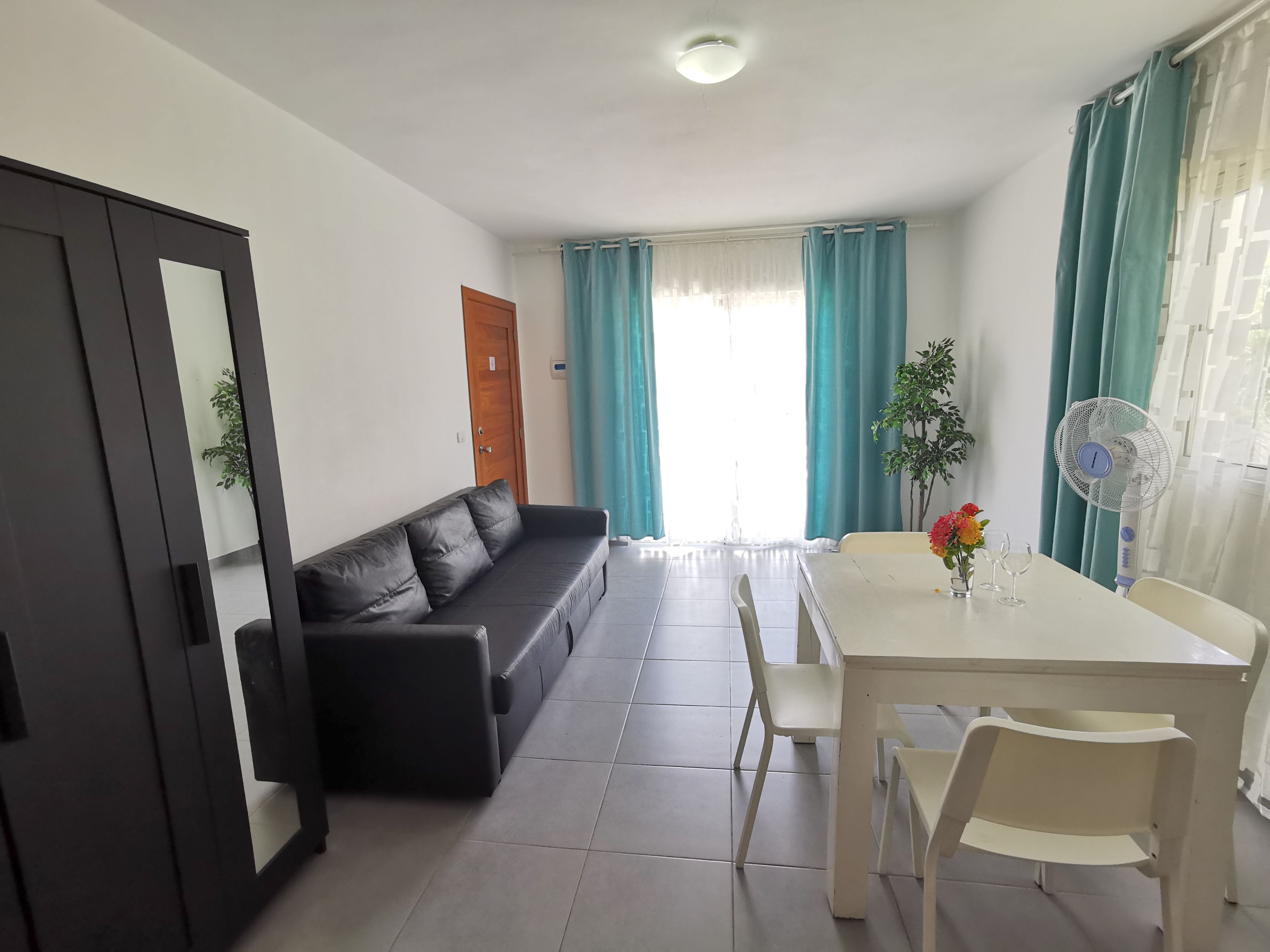 Quarto com sofá de couro preto, mesa de jantar branca com cadeiras, ventoinha no teto e janela com cortinas.