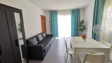 Quarto com sofá de couro preto, mesa de jantar branca com cadeiras, ventoinha no teto e janela com cortinas.