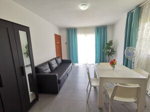 Une pièce avec un canapé en cuir noir, une table à manger blanche avec des chaises, un ventilateur de plafond et une fenêtre avec des rideaux.
