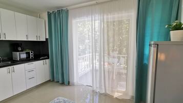 Una cocina con armarios blancos, un microondas y un refrigerador. Hay una ventana con cortinas turquesa y vistas al exterior.