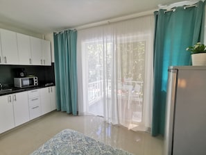 A kitchen with white cabinets, a microwave, and a refrigerator. There is a window with teal curtains and a view of the outdoors.