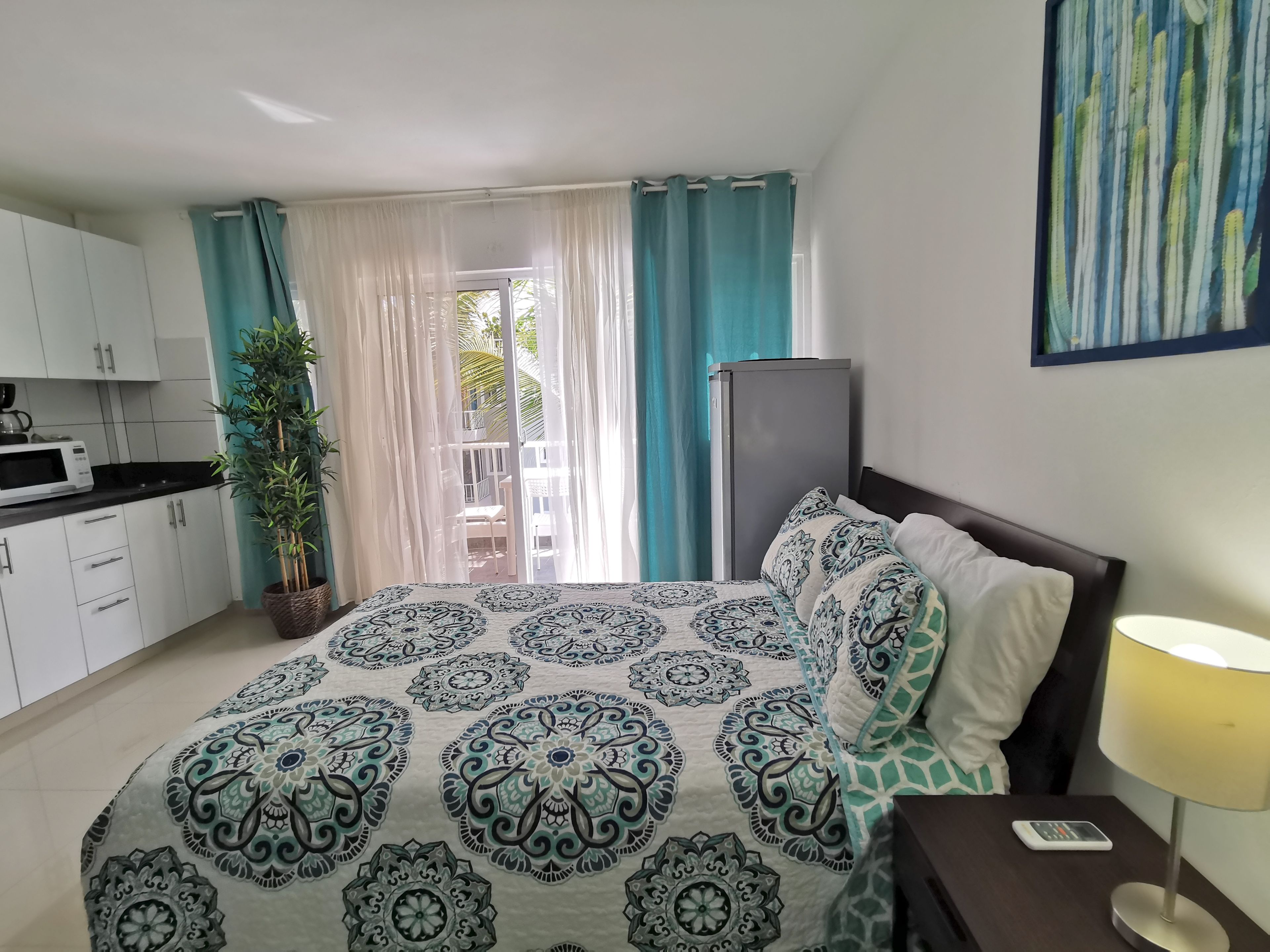 Quarto com cama, mesa de cabeceira com luminária, um aparador, janela com cortinas, vista para o exterior e uma cozinha compacta com micro-ondas.