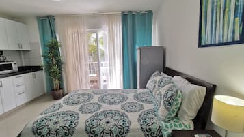 Une chambre avec un lit, une table de chevet avec une lampe, une commode, une fenêtre avec des rideaux, une vue sur l’extérieur et une kitchenette équipée d’un four à micro-ondes.