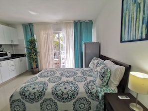 Un dormitorio con cama, mesita de noche con lámpara, ventana con cortinas, vistas al exterior y una kitchenette con microondas.