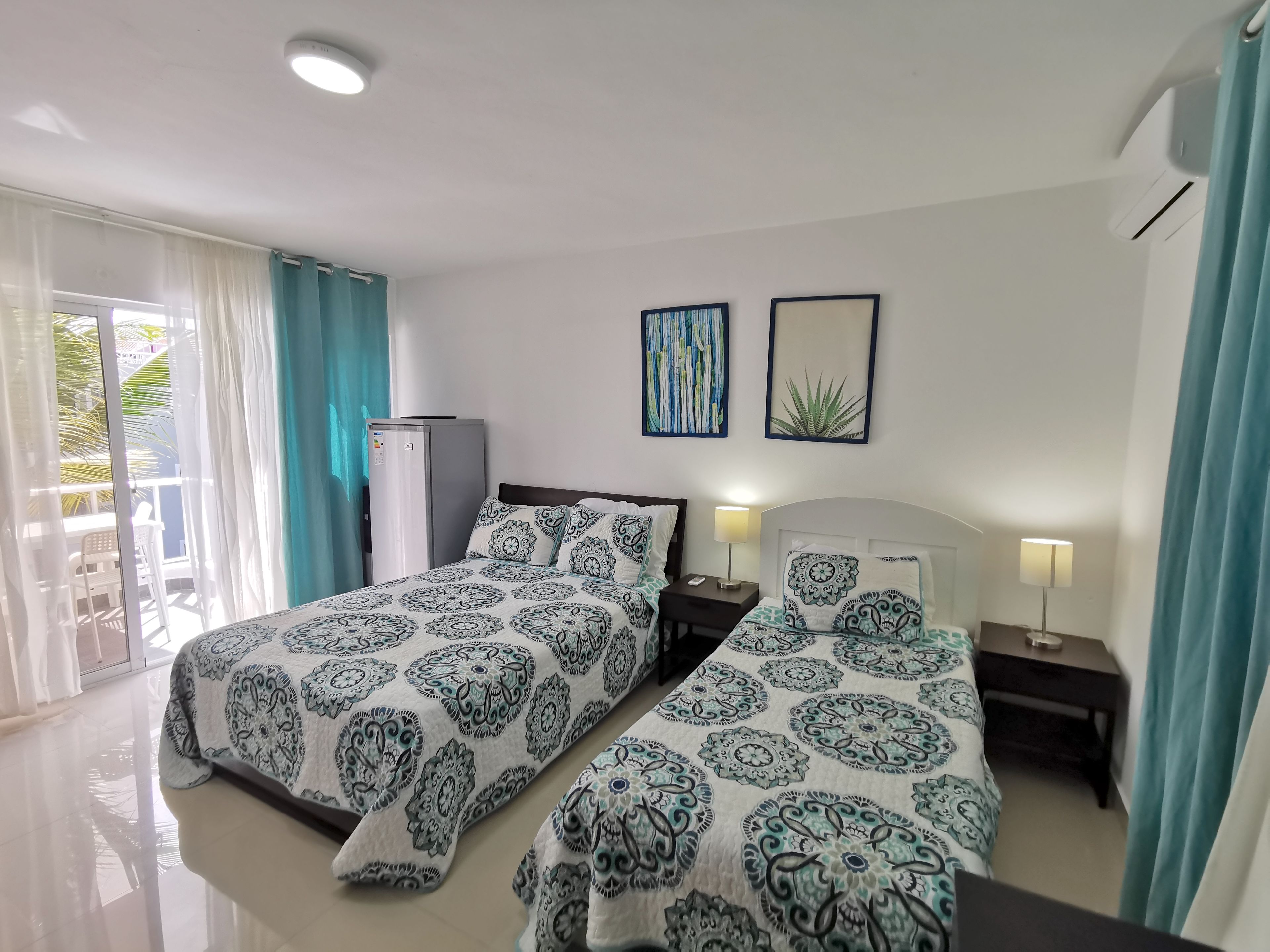 A hotel room with two beds, a refrigerator, a nightstand with a lamp, and a window with curtains.
