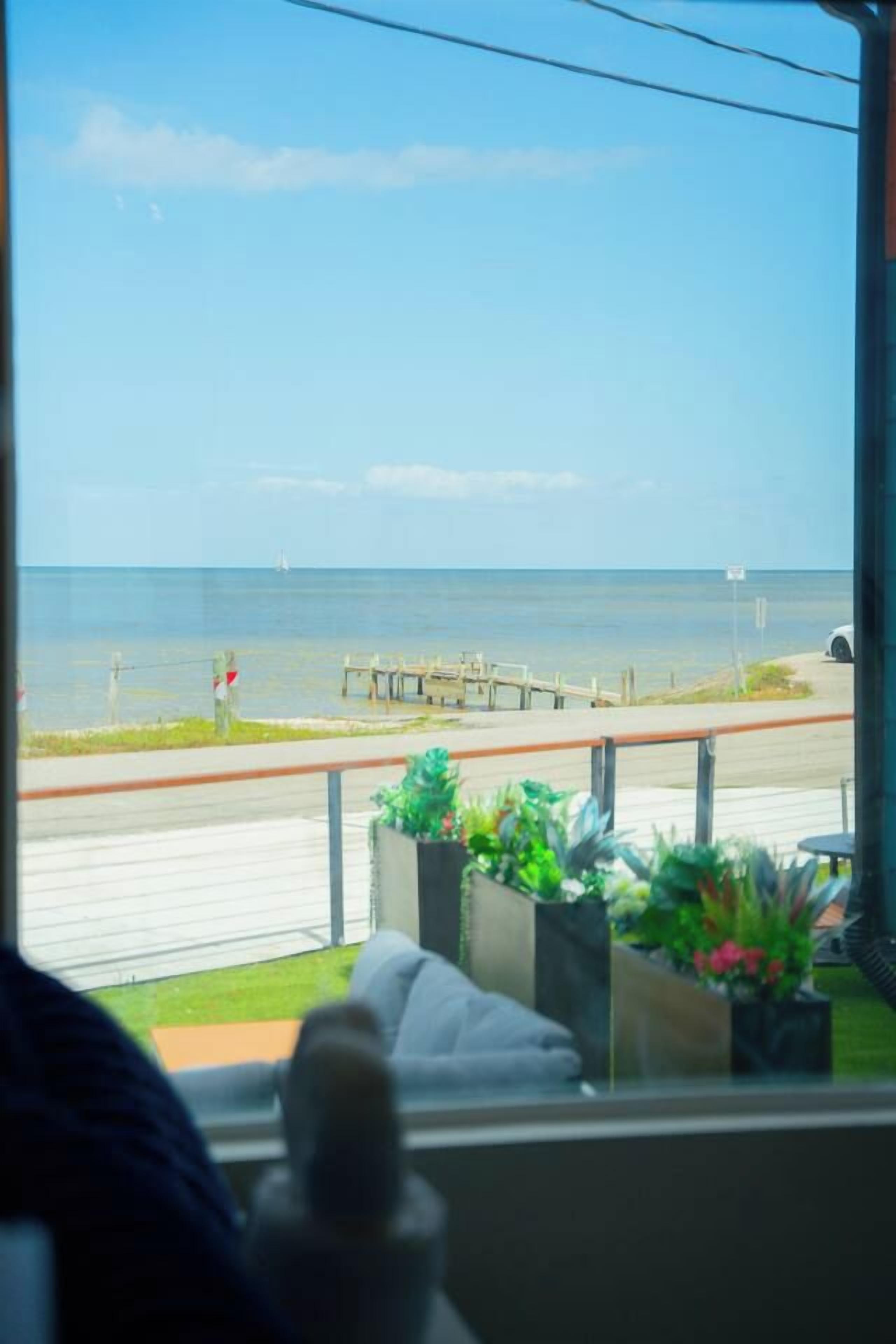 Uitzicht vanuit een raam op een strand met een houten pier, een weg en een geparkeerde auto. Op de vensterbank staan potplanten.