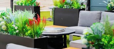 An outdoor seating area with grey sofas and a round table, surrounded by potted plants.