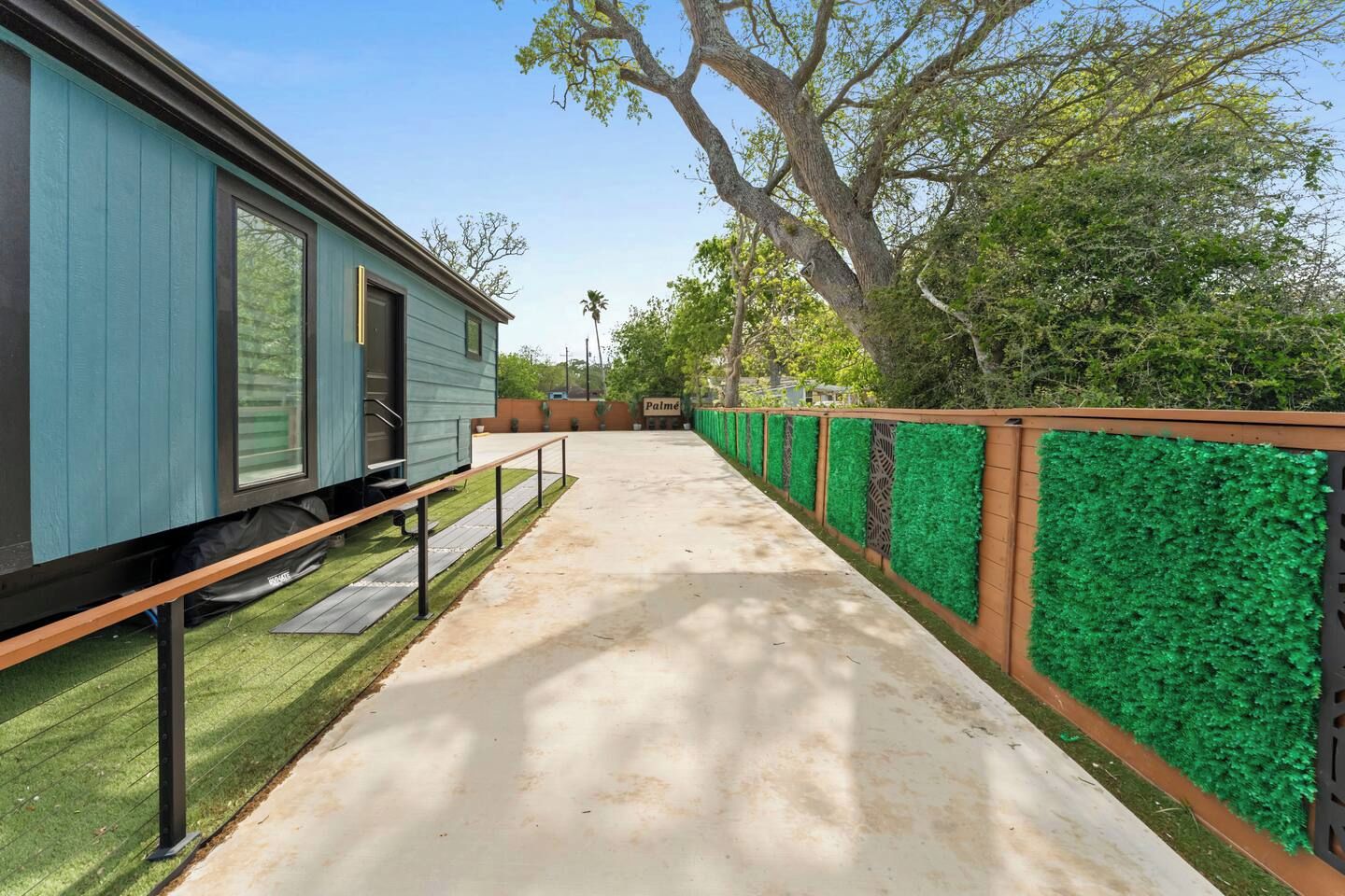Un sendero conduce a un edificio con fachada azul, rodeado de césped artificial verde y una valla de madera.