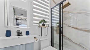 Et moderne badeværelse med en hvid kommode, toilet, brusekabine i glas og en lille potteplante på kommoden.