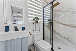 A modern bathroom with a white vanity, a toilet, a glass shower enclosure, and a small potted plant on the vanity. - Tiny Lux Home | Pool + Bayfront Views | Near Kemah (Bacliff)