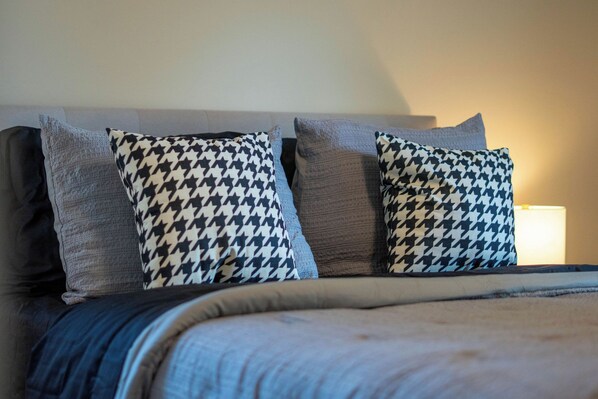 Una cama con cabecera gris, dos almohadones estampados y una lámpara de noche.