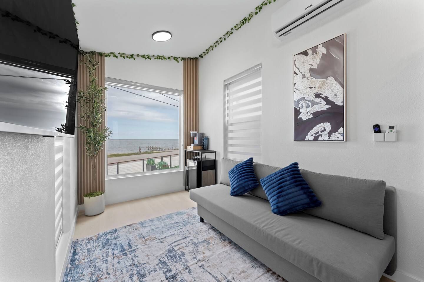 Una sala de estar moderna con un sofá gris, cojines azules, un ventanal con vista al mar, una planta en maceta y un aire acondicionado de pared.