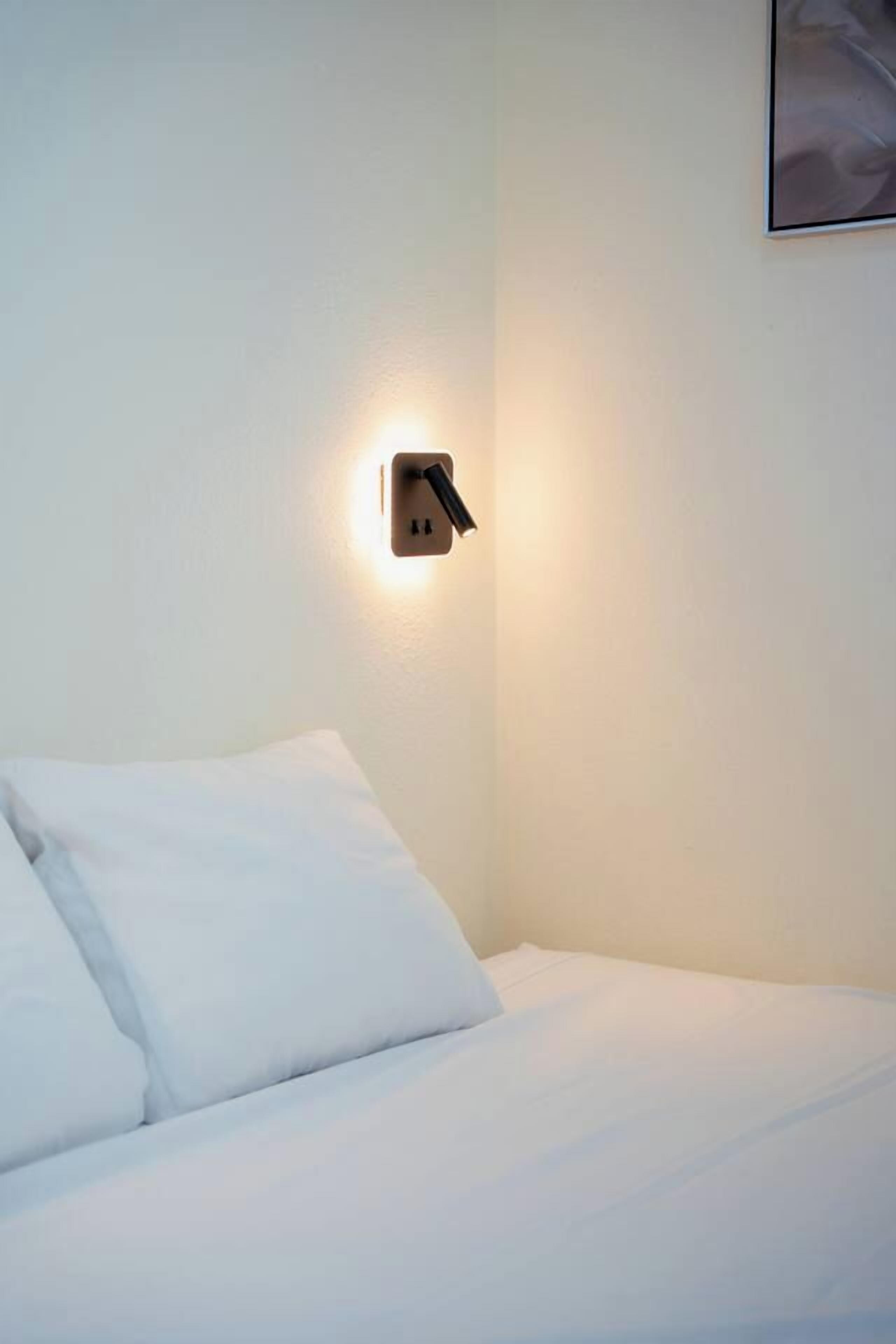 A hotel room with a neatly made bed, a wall-mounted light fixture, and a framed picture on the wall.